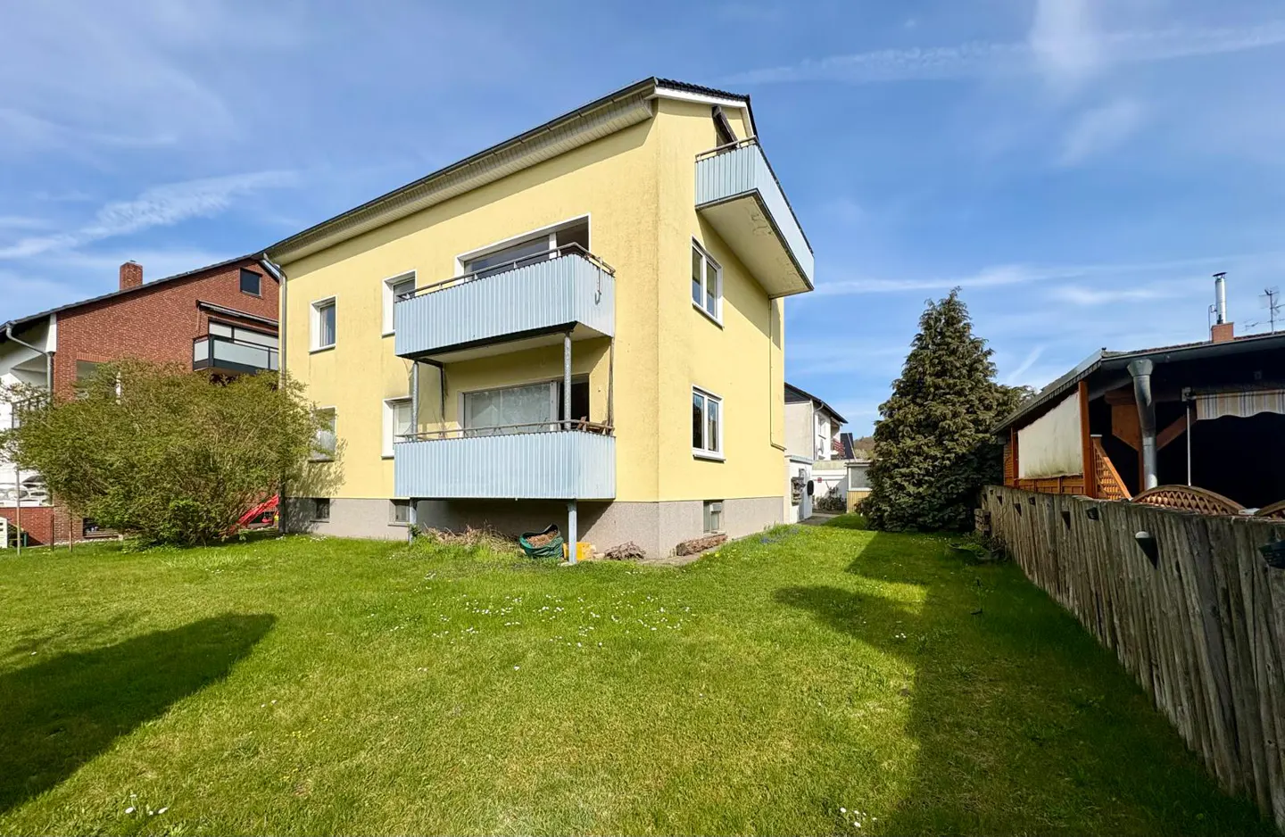 Exterior view of a yellow two-story house with blue balconies, surrounded by a green lawn and a wooden fence.