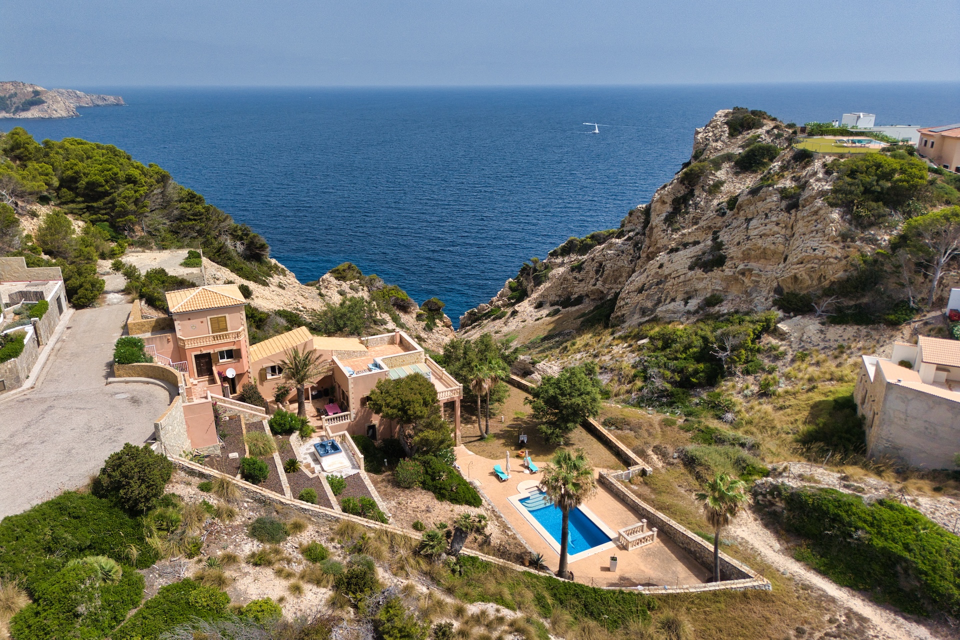 Aerial view of a peach-colored villa with a pool, nestled in a rocky coastal landscape overlooking the blue Mediterranean.