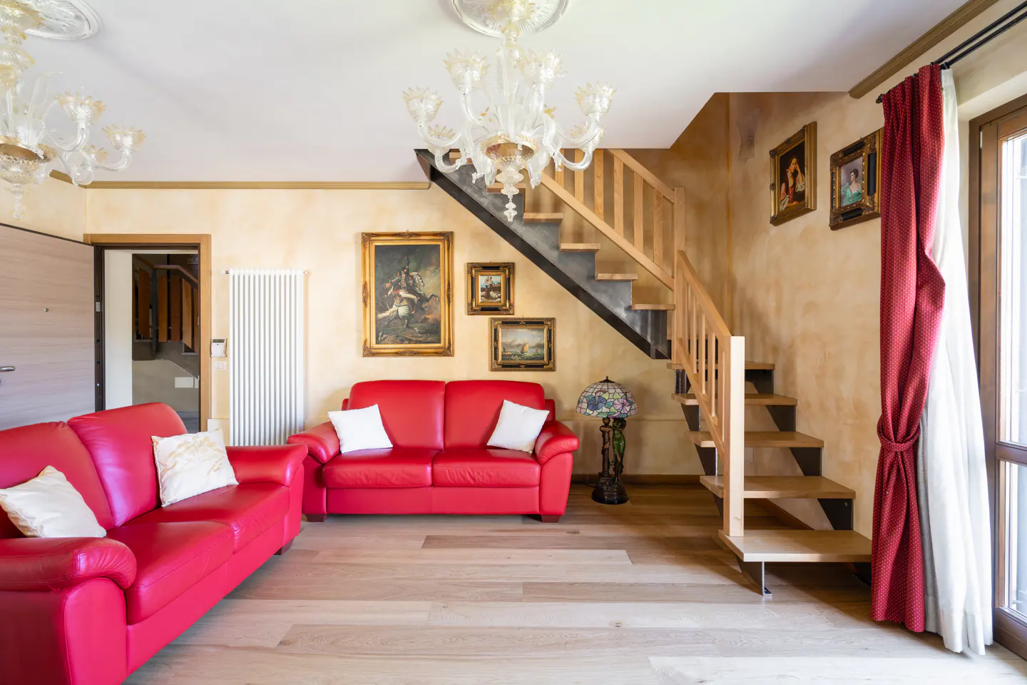 Living room with red sofas, wood floors, and a staircase. Paintings adorn the walls, and a chandelier hangs from the ceiling.