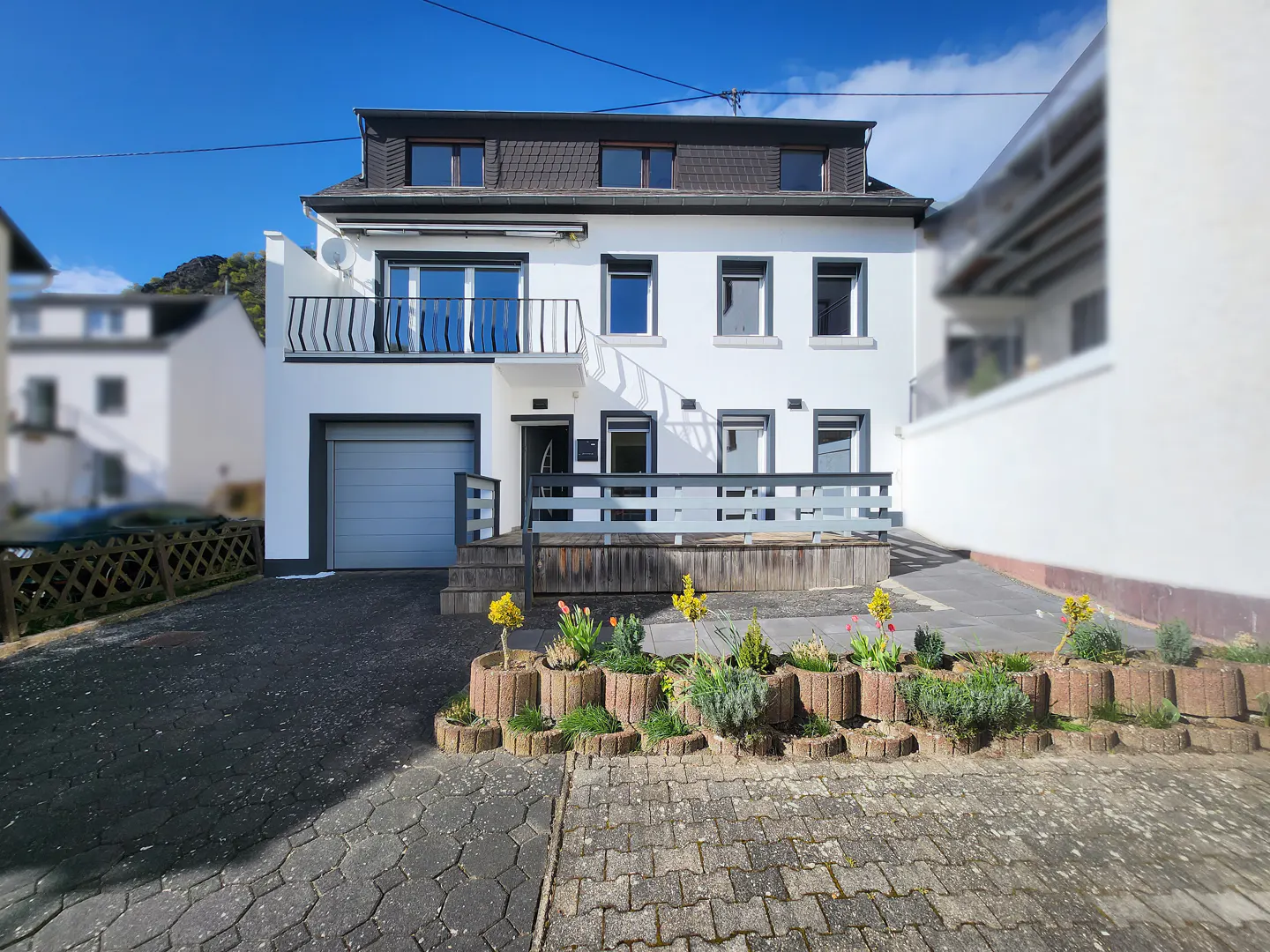 A three-story white house with a gray garage door and a small balcony. Flower beds line the front of the house.