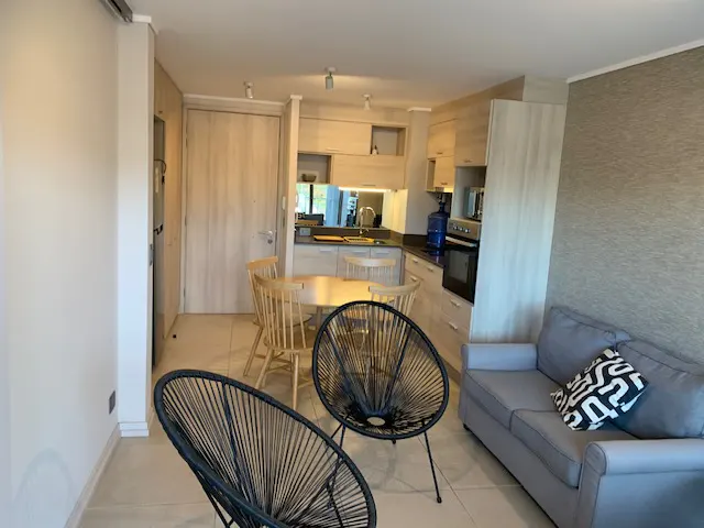 Interior view of a living space with a gray sofa, black chairs, and a round wooden table near a kitchen area with light wood cabinets.