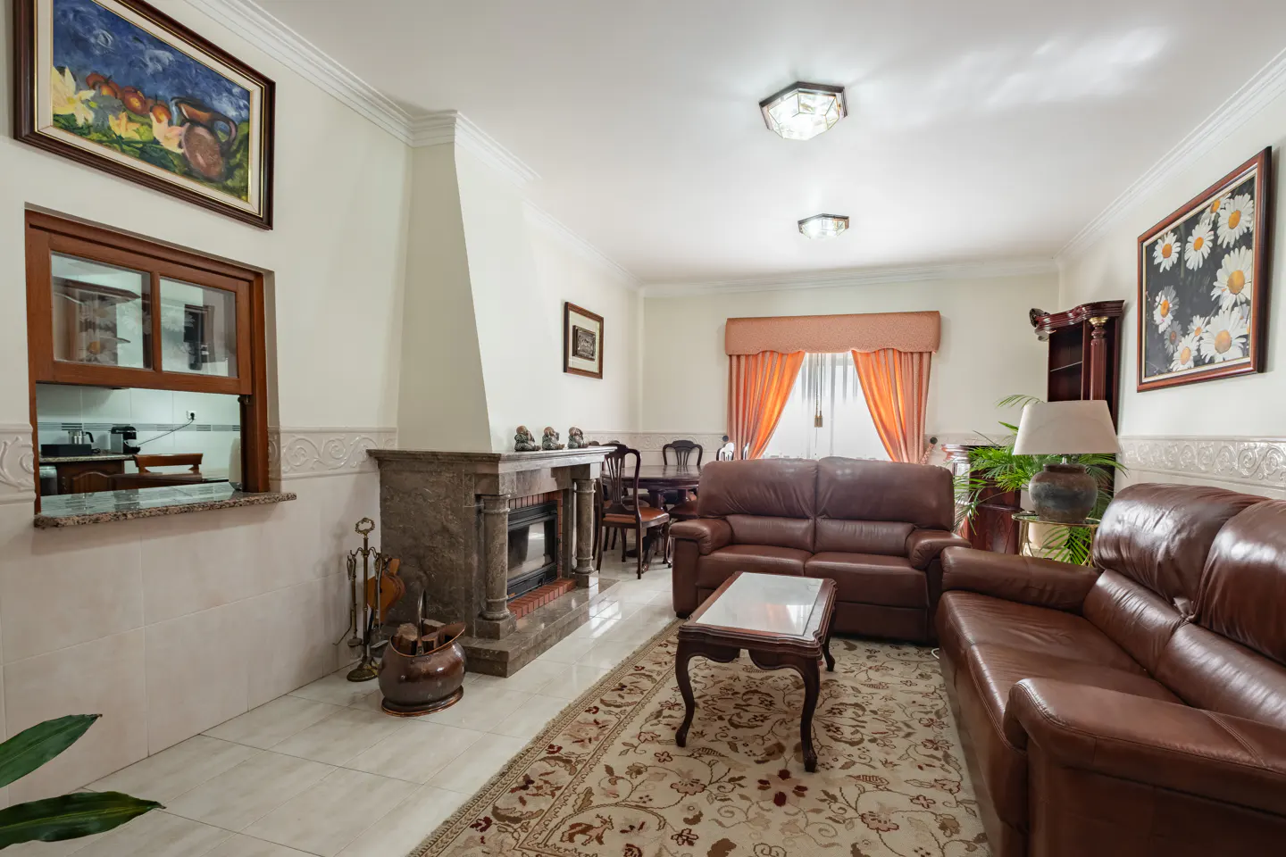 A living room with brown leather sofas, a marble fireplace, and a floral rug. A dining table is visible in the background.