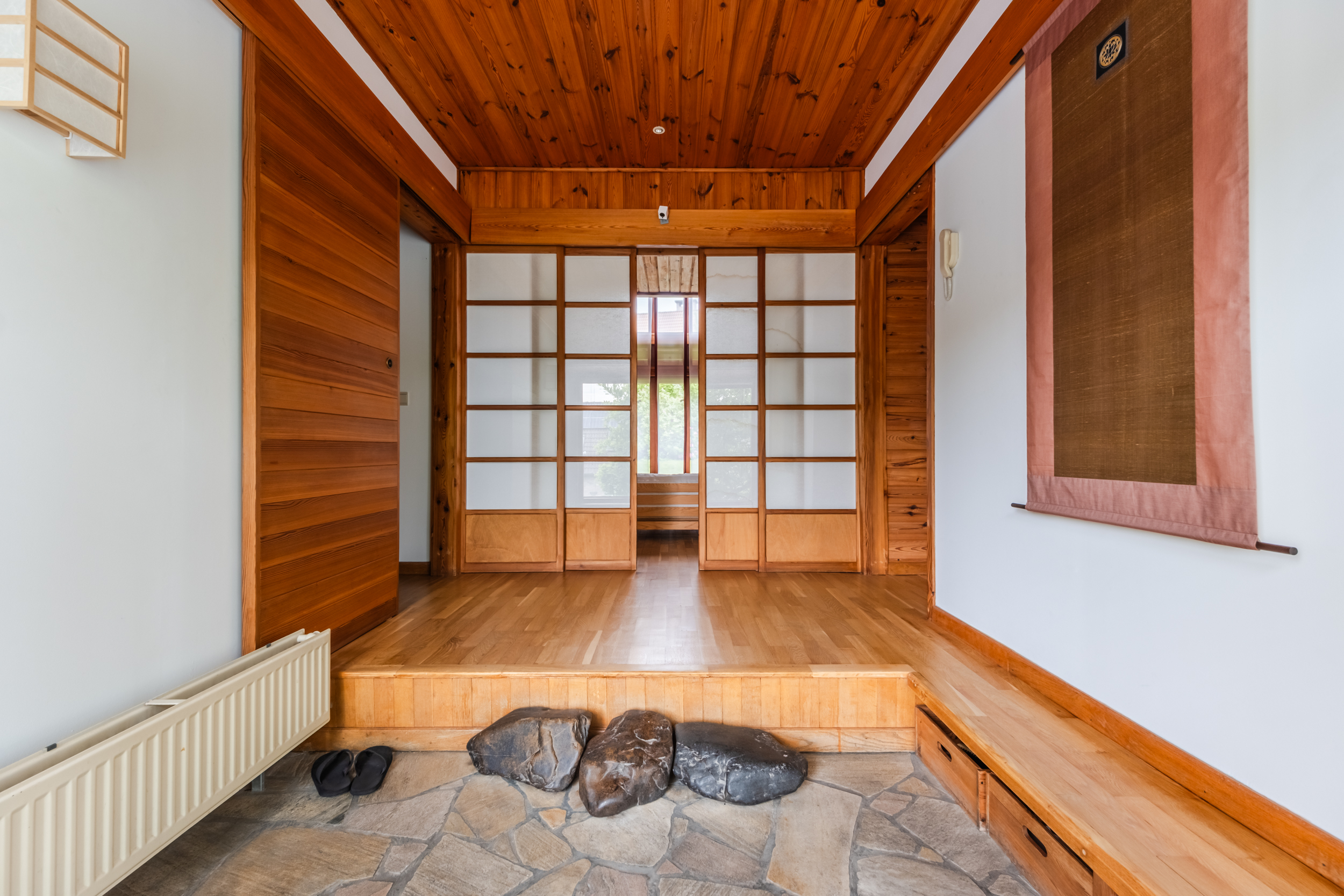 Interior view of a Japanese-style home with wood floors, shoji screens, and a stone floor entrance. A scroll hangs on the white wall.