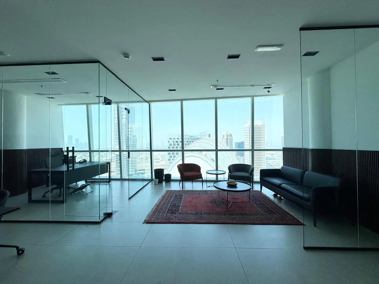 Bright office space with glass walls, a black sofa, and a red rug. City view through large windows.