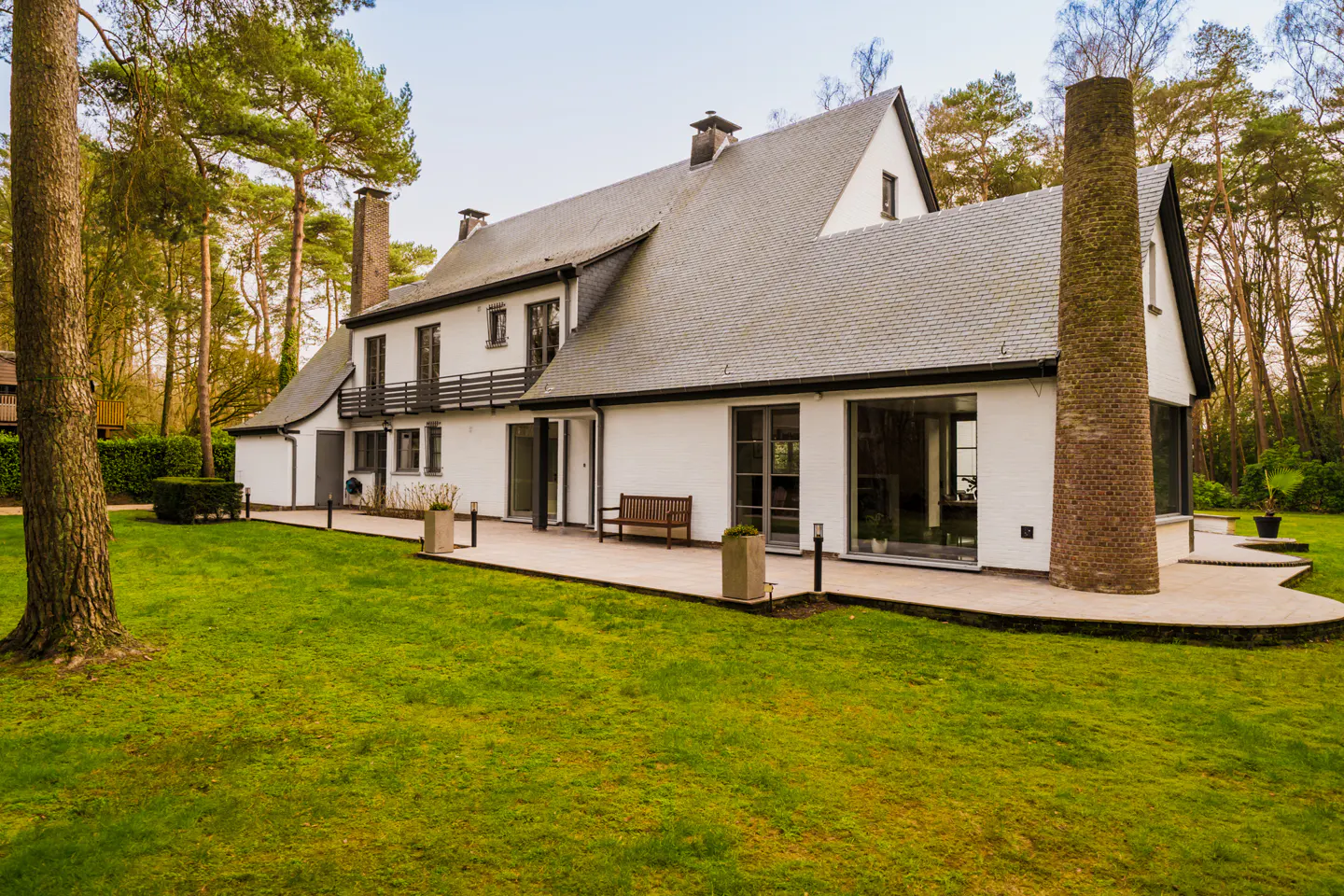 Exterior view of a large white house with a gray roof, surrounded by green lawn and trees. A patio with a bench extends from the house.