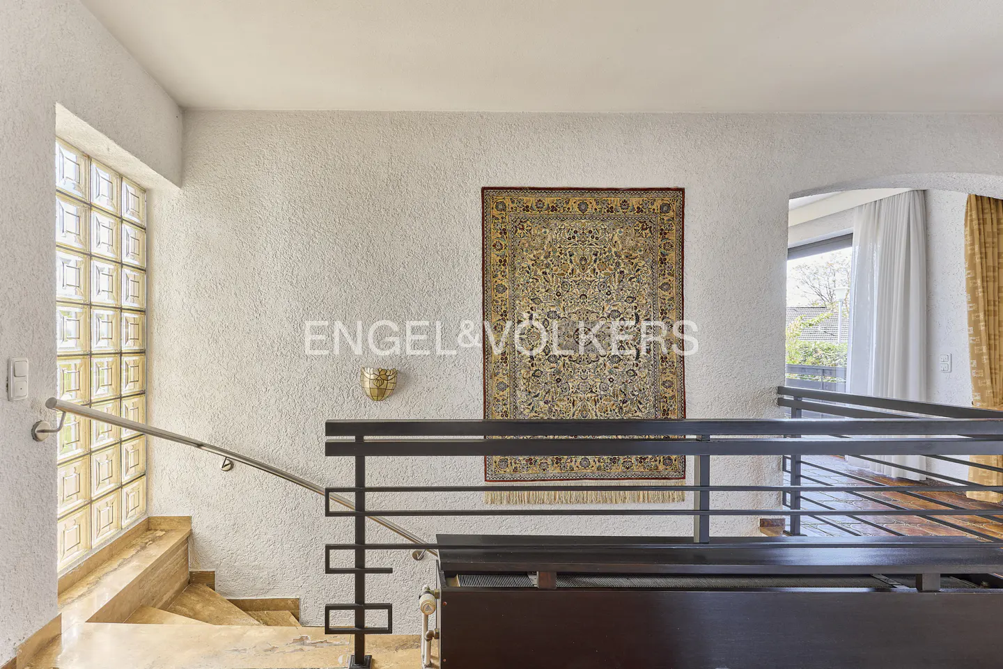 Interior view of a staircase with marble steps, a metal railing, and a decorative rug hanging on the wall.