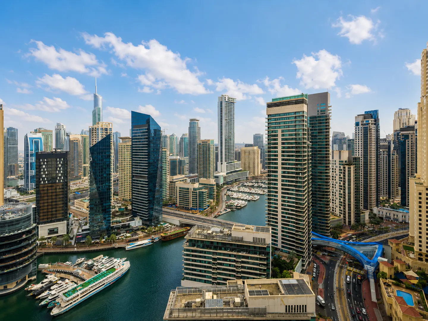 Dubai Marina cityscape view with skyscrapers, boats, and a blue sky with white clouds.