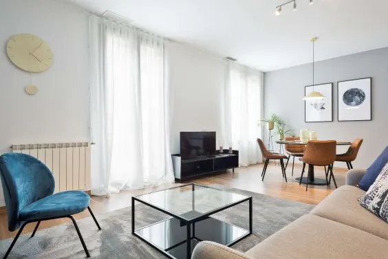 Modern living room with a blue armchair, glass coffee table, beige sofa, and a dining area with brown chairs and art.