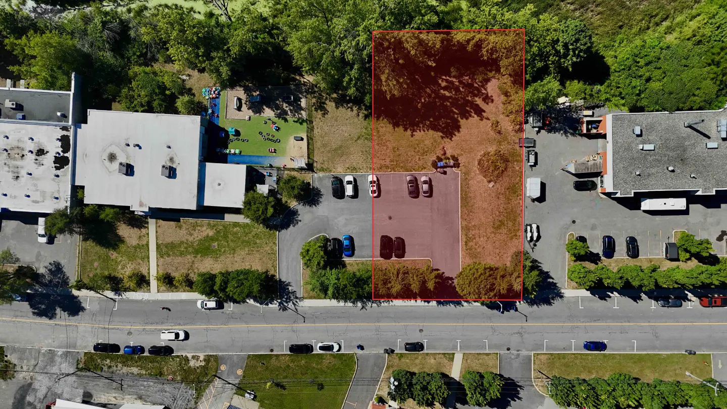 Aerial view of a rectangular property outlined in red, adjacent to a parking lot and buildings. Trees surround the lot.