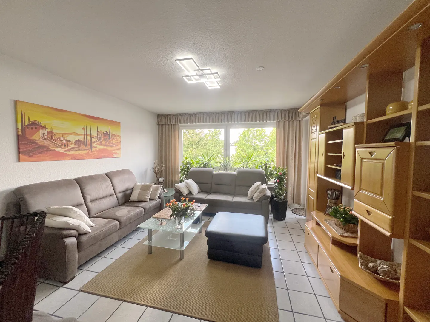 Living room with brown sofas, glass table, ottoman, and light wood built-in shelves. A landscape painting hangs on the wall.