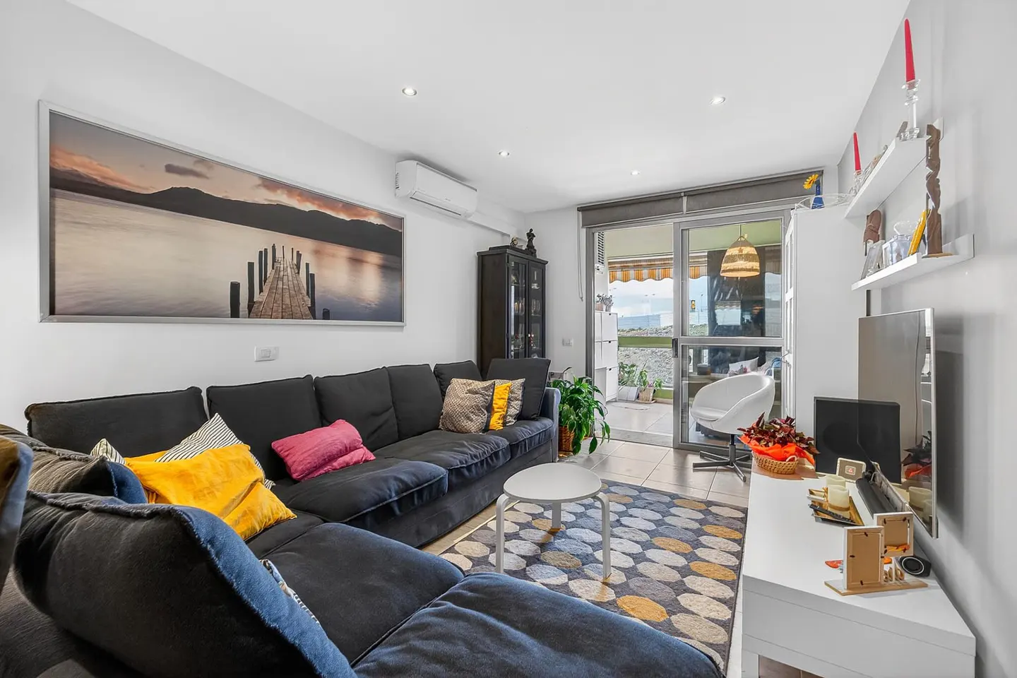 Bright living room with a large gray sectional sofa, colorful pillows, and a lake scene wall art. Balcony access and white furniture.
