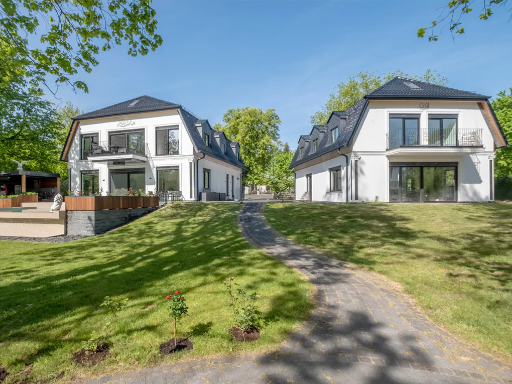 Two modern white houses with black roofs face each other across a green lawn and stone path under a blue sky.
