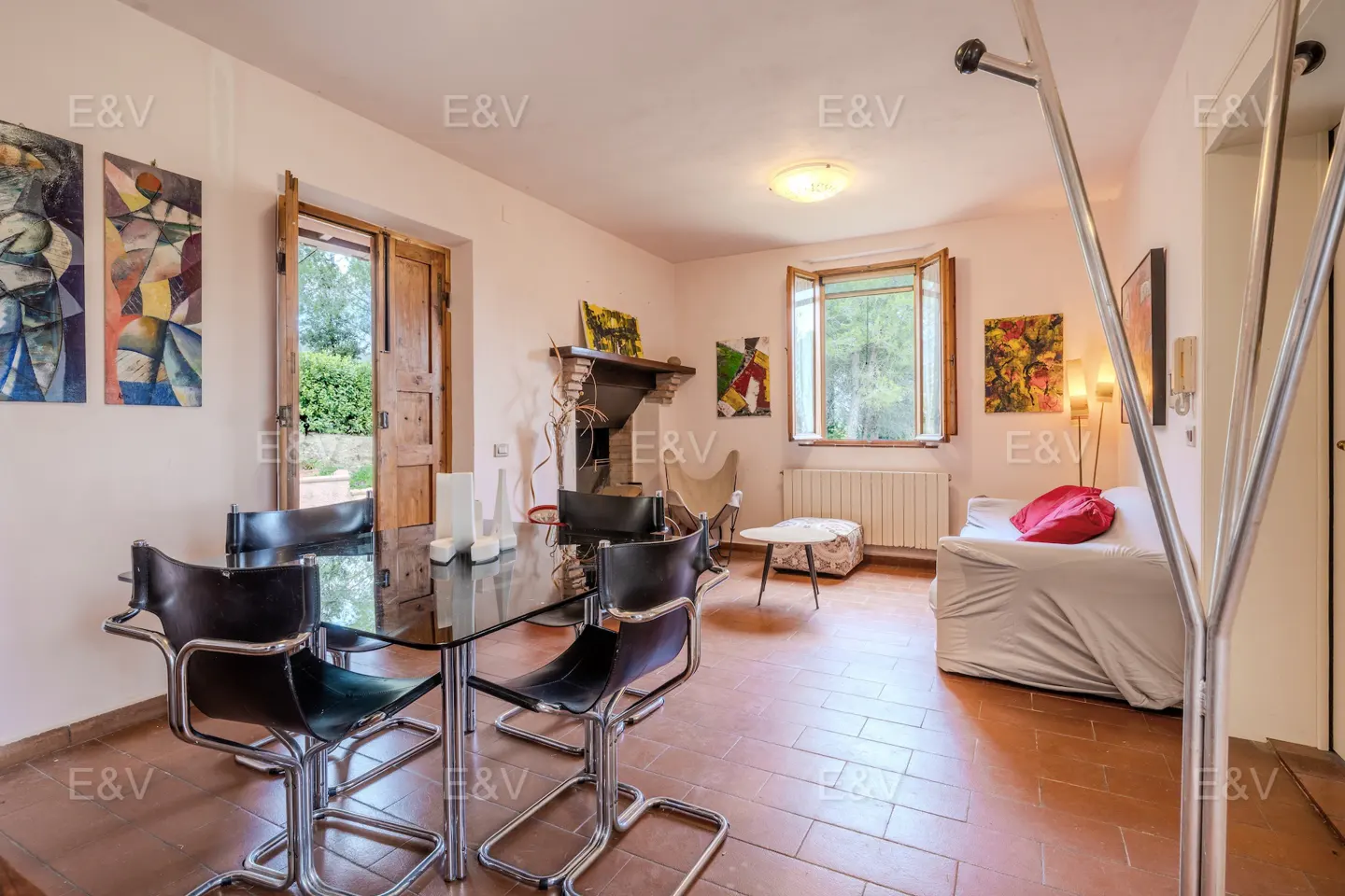 A bright living room with a glass table, black chairs, and a white sofa with a red pillow. Artwork adorns the walls.