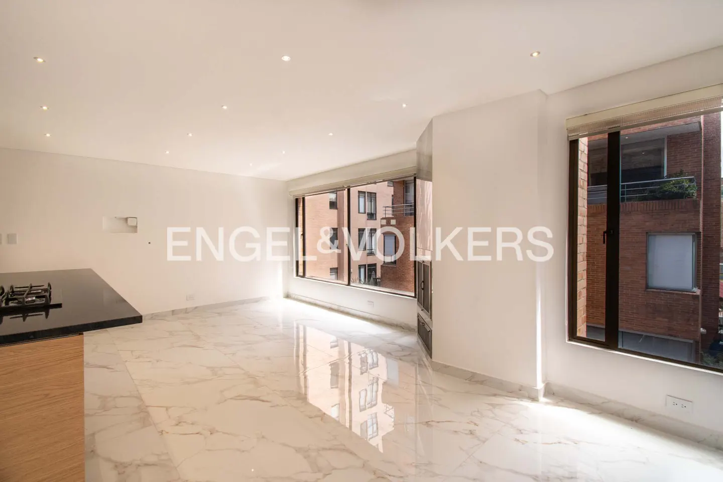 Bright, modern apartment interior with white walls, marble floors, and large windows overlooking a brick building. A black countertop is visible.
