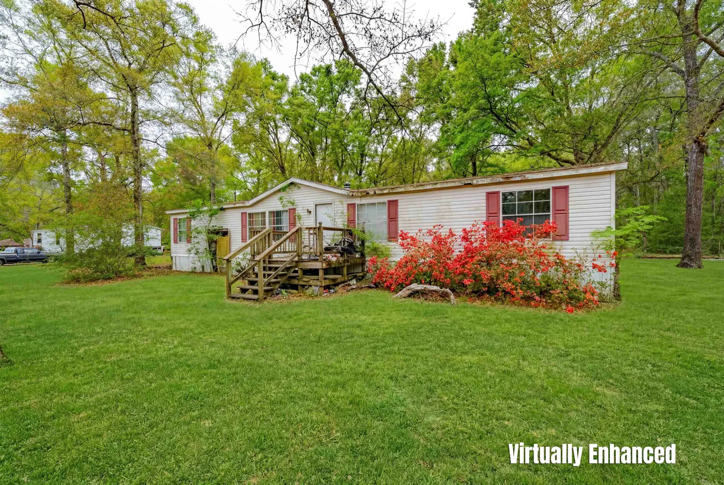 Exterior view of a white mobile home with red shutters, a wooden porch, and red flowers in the front yard. Green trees surround the property.
