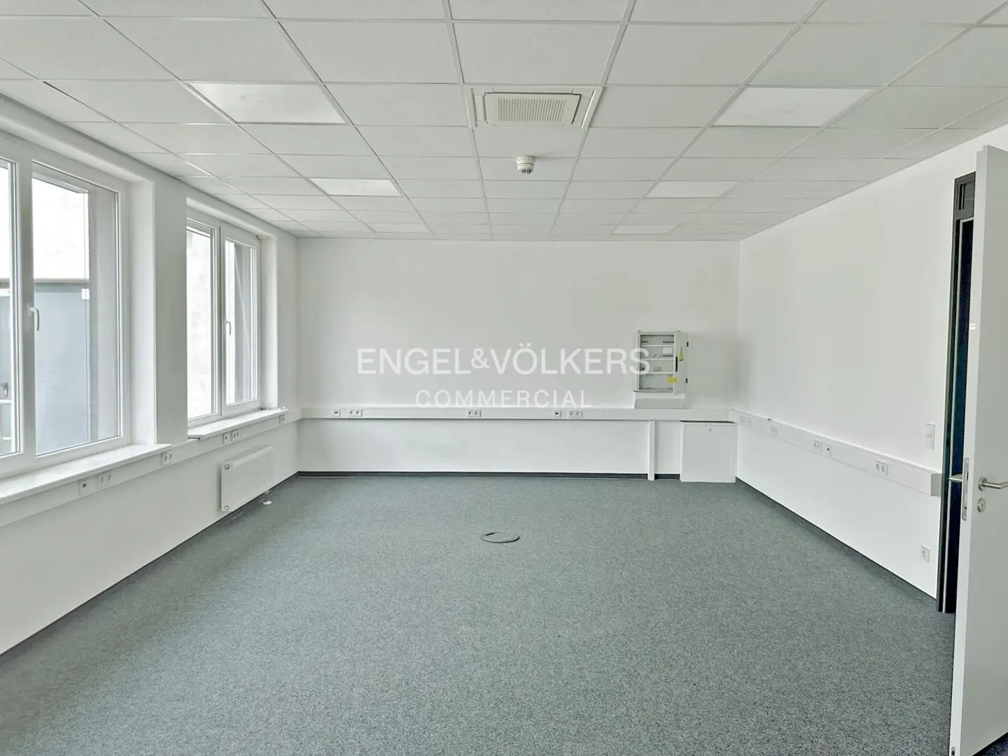 Bright, empty office space with gray carpet, white walls, and windows. Engel & Völkers Commercial logo on the wall.