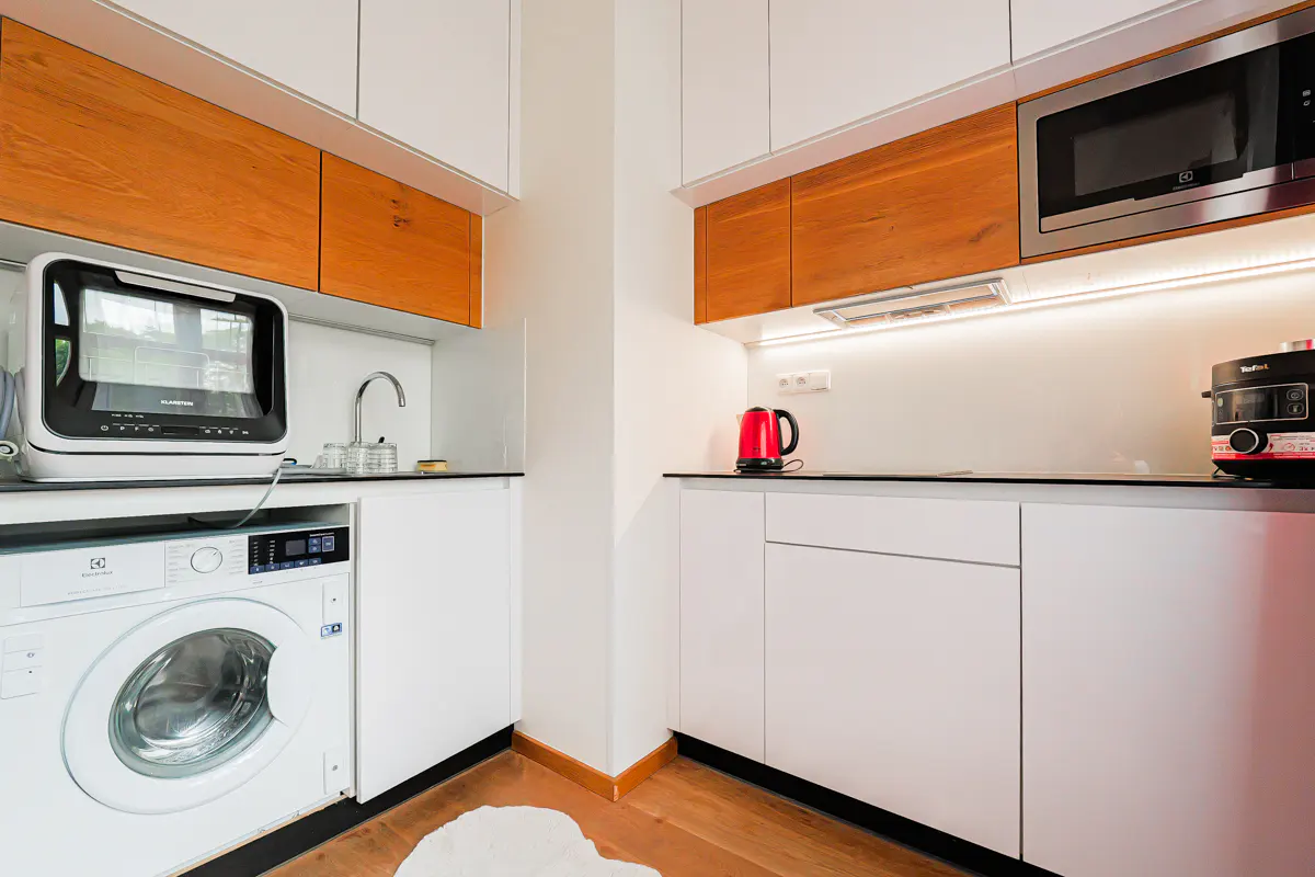 Modern kitchen with white cabinets, wood accents, a washing machine, microwave, and a red kettle on a wooden floor.