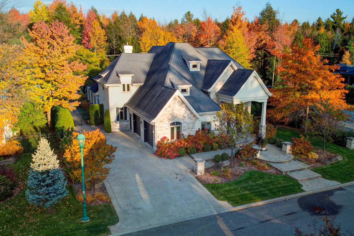 Aerial view of a large, two-story house with a gray roof, surrounded by colorful autumn trees. A long driveway leads to a two-car garage.