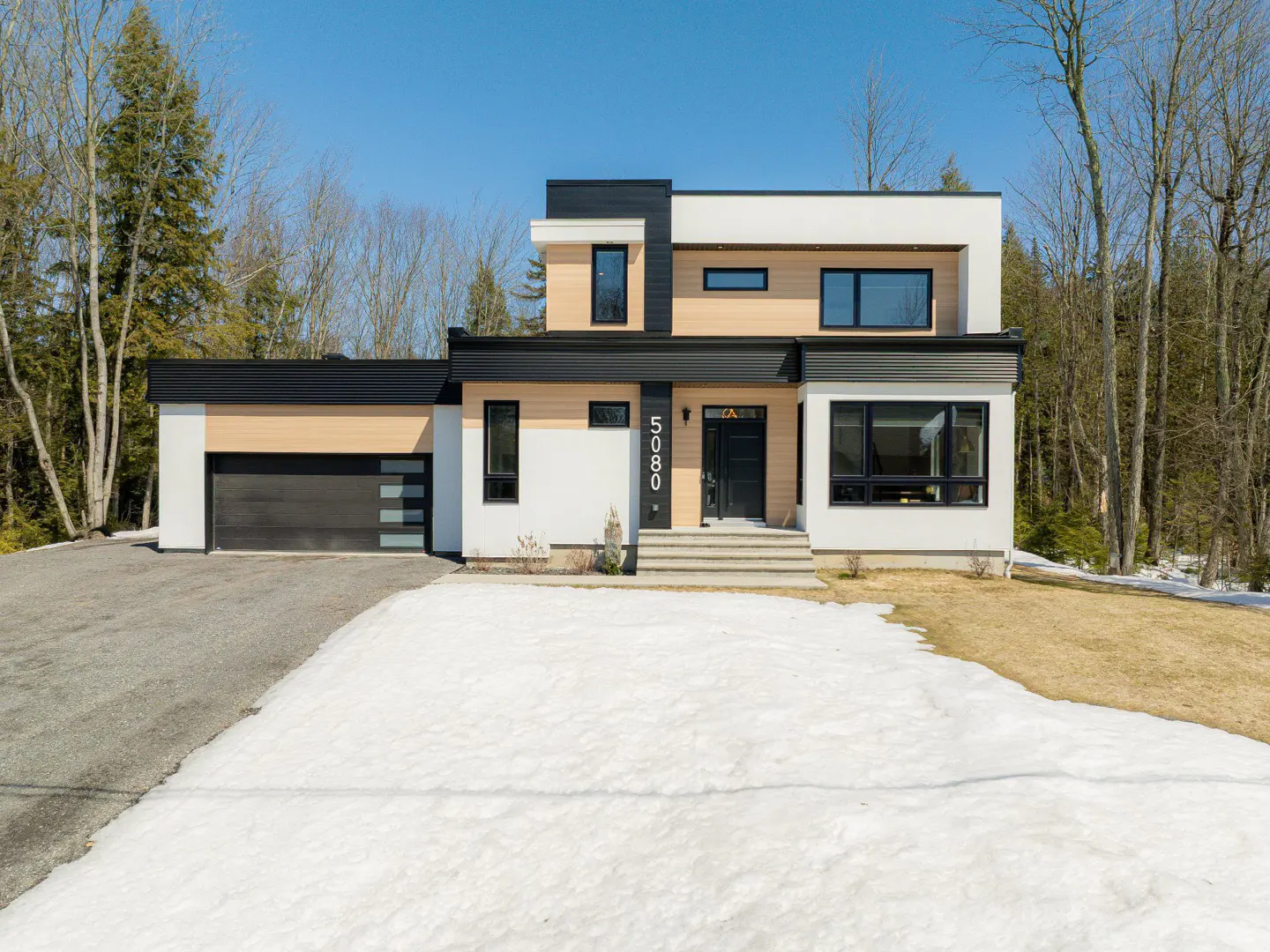 Modern two-story home with black trim, light wood paneling, and a black garage door. Snow covers the front yard.