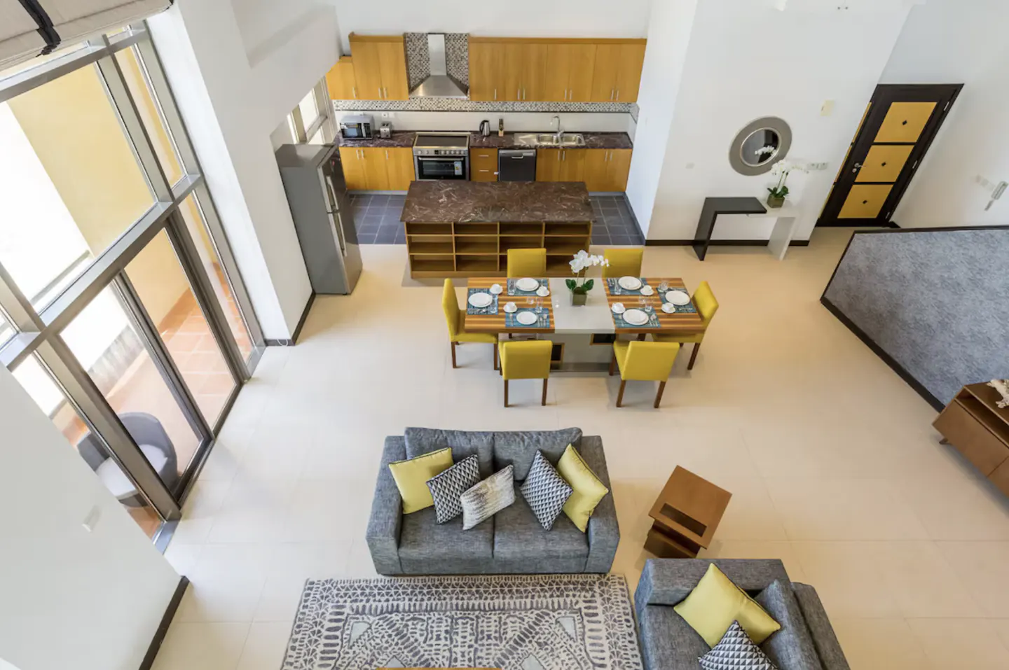 Overhead shot of a modern open-concept living space with a kitchen, dining area, and lounge with gray sofas and yellow accents.