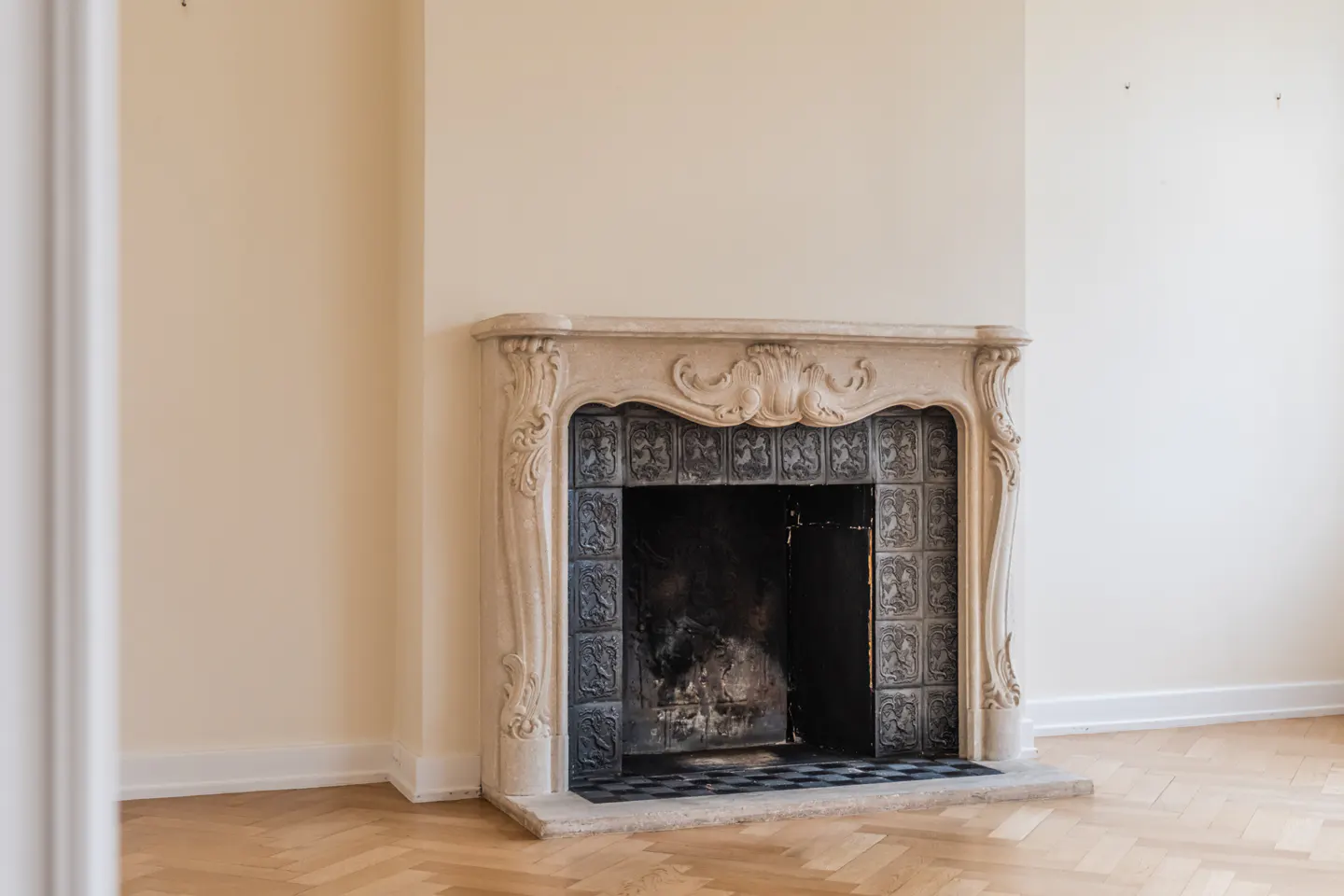 Ornate fireplace with a light stone mantel and decorative tiles, set against a cream-colored wall and herringbone wood floor.
