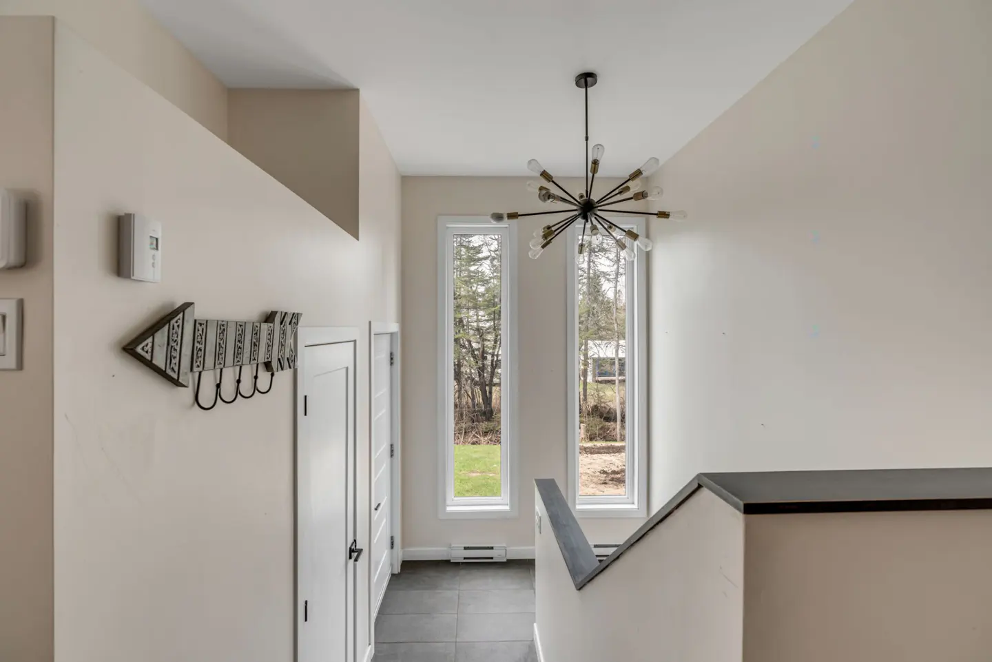 Interior view of a foyer with white walls, dark tile floor, and a modern starburst chandelier. Two tall windows overlook a green lawn and trees. An arrow-shaped coat rack hangs on the wall.