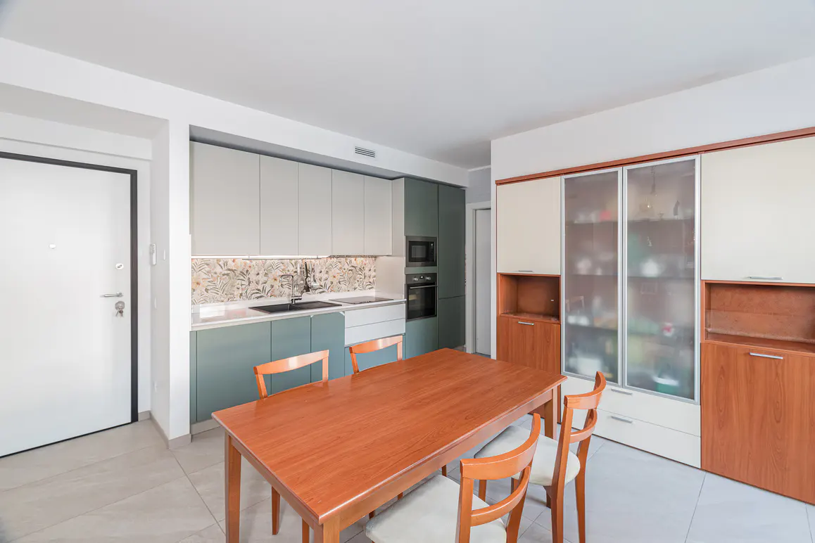 A modern kitchen with a wooden table and chairs in the foreground. Cabinets are two-toned, with floral backsplash. A glass-fronted cabinet is on the right.