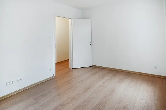 Empty room with light wood floors, white walls, and an open white door leading to another room.