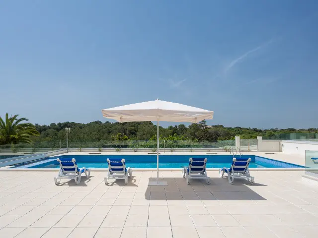 A sunny poolside view with four blue and white lounge chairs under a large white umbrella, overlooking green trees.
