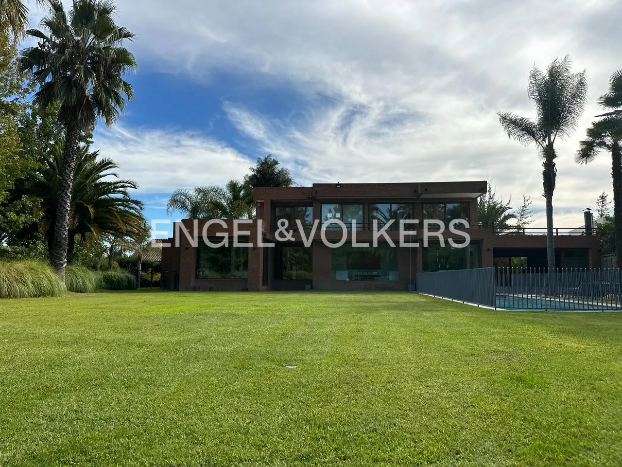 Modern brown house with large windows, surrounded by green lawn and palm trees under a cloudy blue sky. Engel & Volkers logo visible.