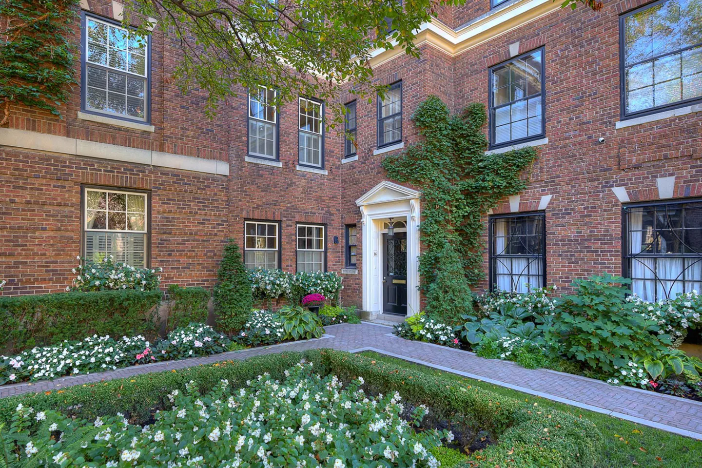 Brick building with black-framed windows and a black door, surrounded by a garden with white flowers and green bushes.