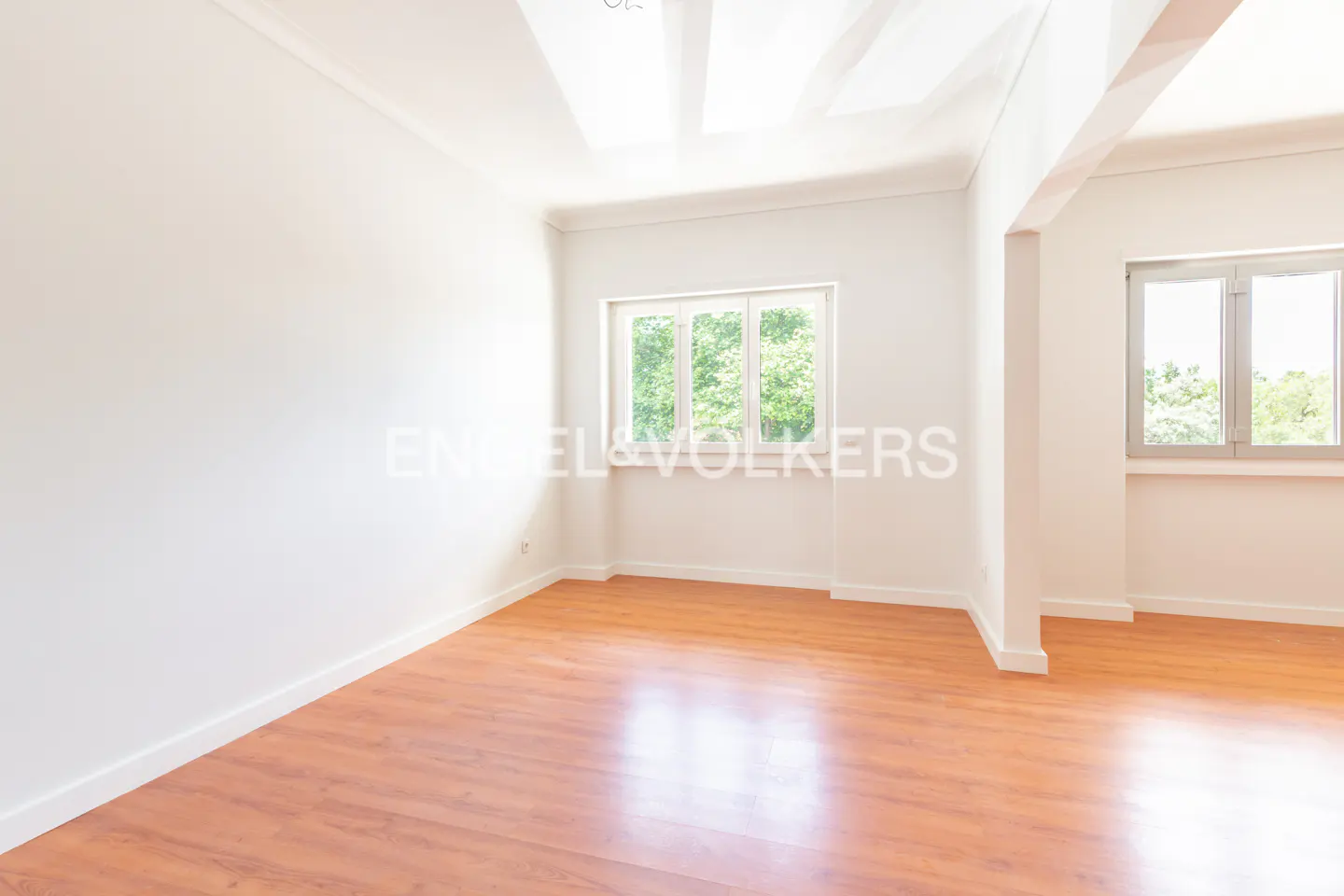 Bright, empty room with wood floors, white walls, and two windows showing green trees outside.