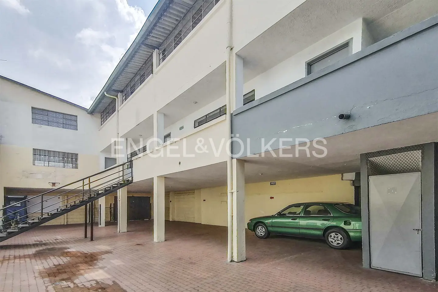 Exterior view of a multi-story building with a green car parked underneath, a staircase, and a brick courtyard.