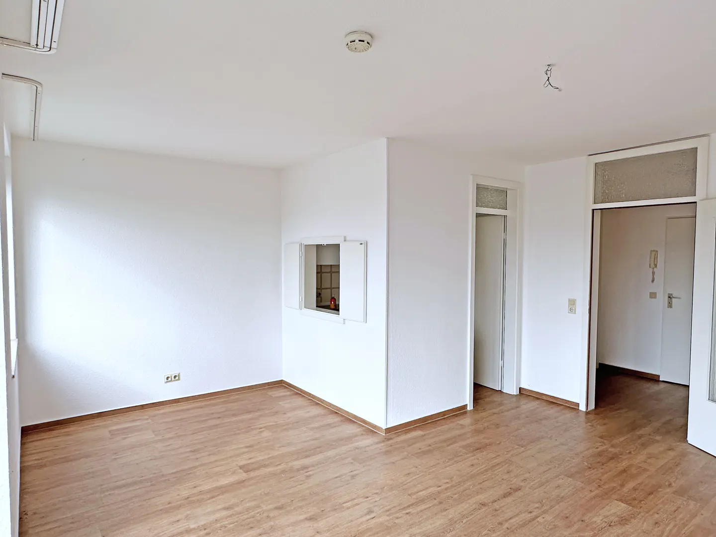 Bright, empty room with wood floors and white walls. A pass-through window looks into the kitchen. Two doorways lead to other rooms.