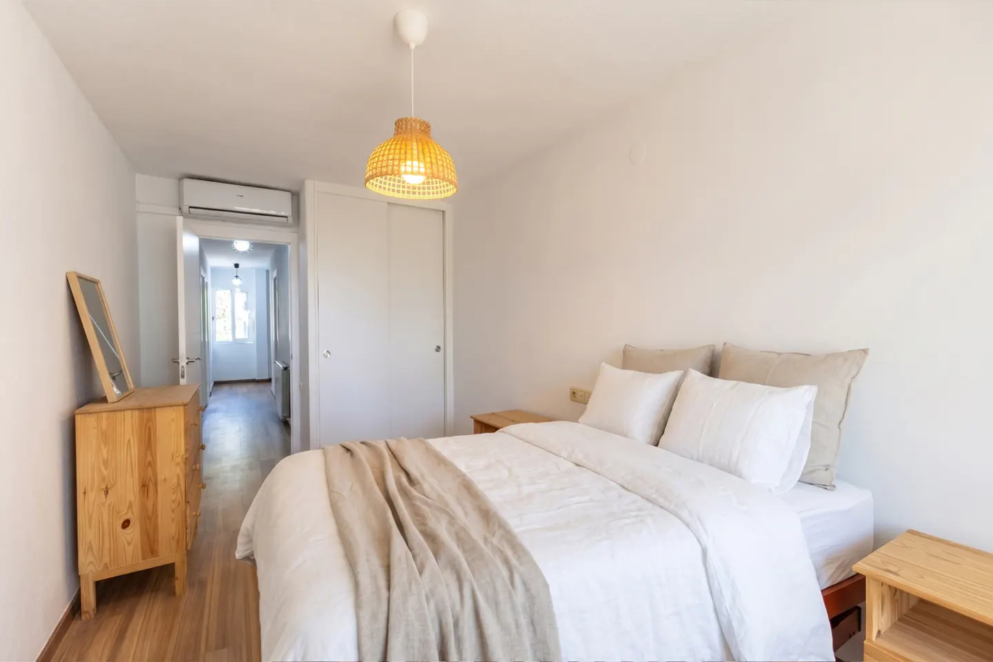 Bright bedroom with white walls, a bed with white linens and a beige throw, and wooden furniture. A hallway is visible in the background.
