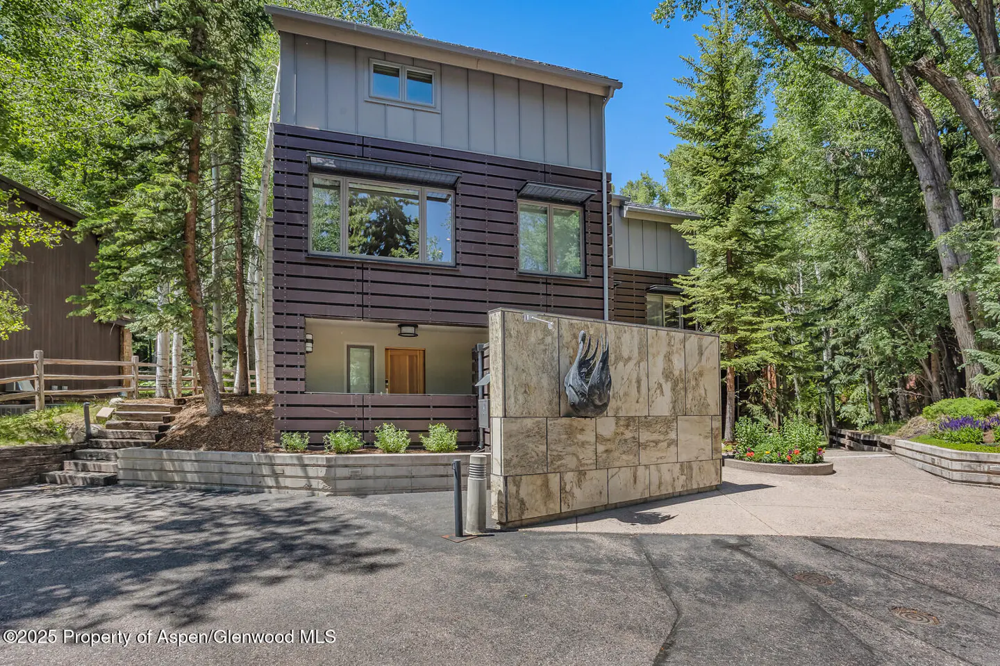 Modern two-story home with brown wood and gray siding, surrounded by trees. A stone wall with a swan sculpture stands near the driveway.