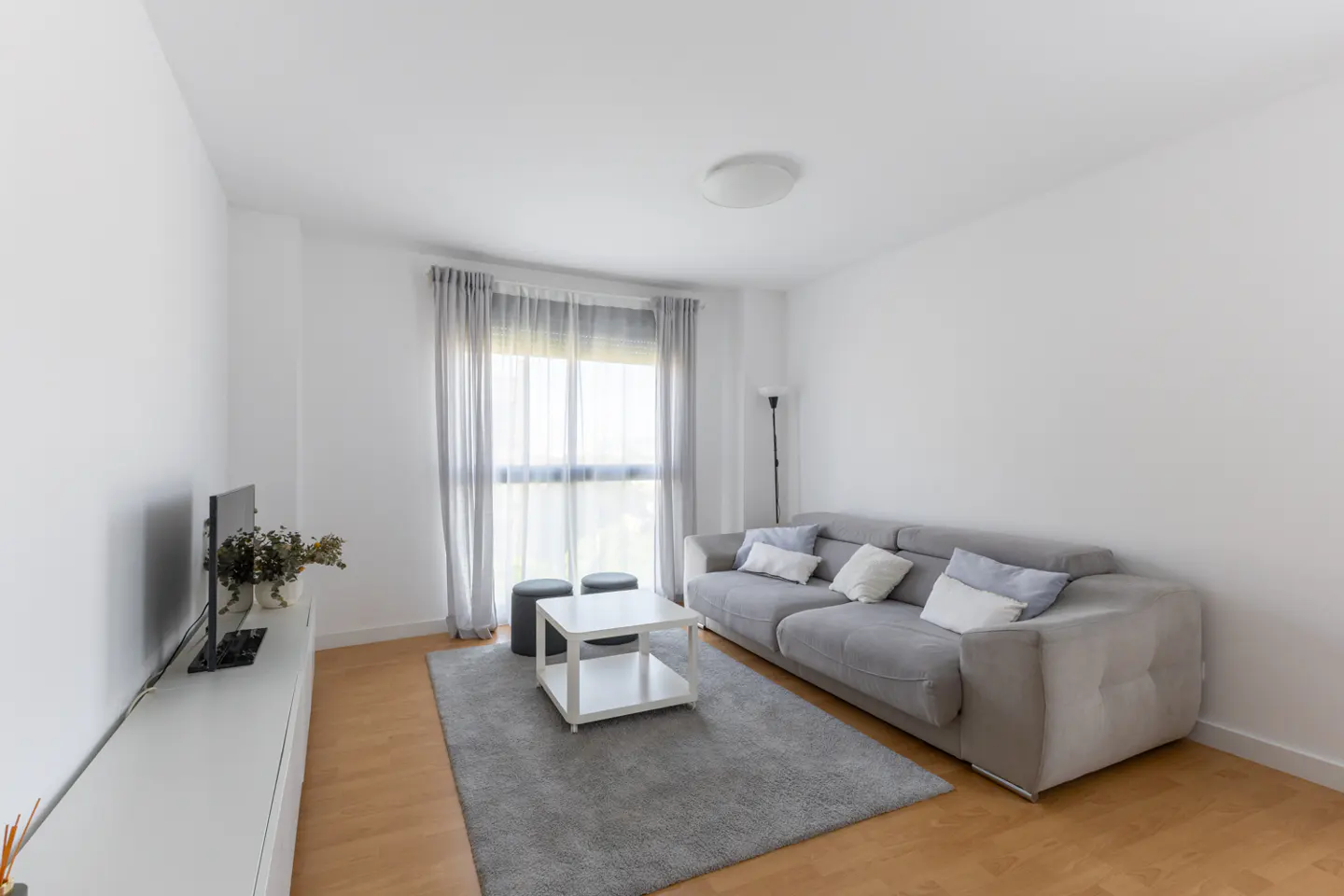 Bright living room with gray sofa, white walls, wood floors, and a gray rug. A white table sits in the center.
