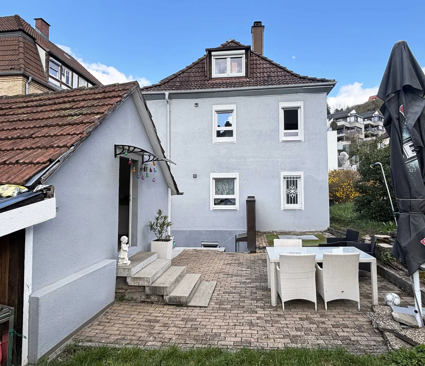 Exterior view of a two-story gray house with a red tile roof and a brick patio with white outdoor furniture.