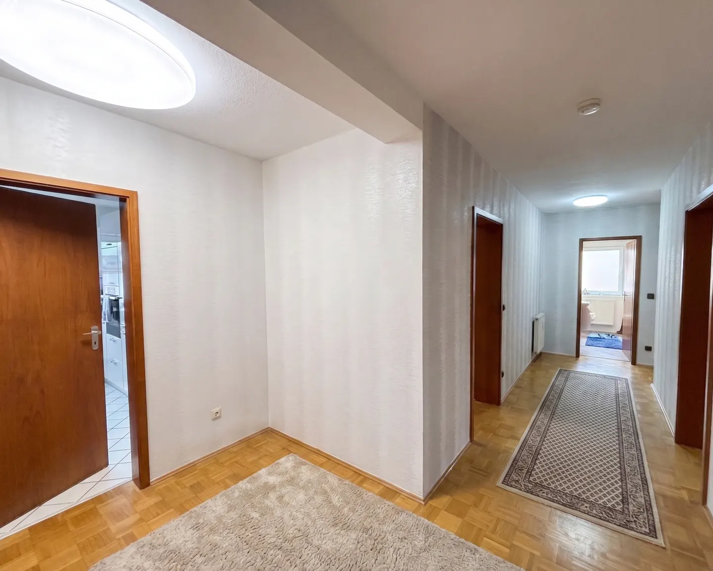 Hallway with wood floors, white walls, and brown doors. A beige rug is on the floor. A kitchen is visible through an open door.