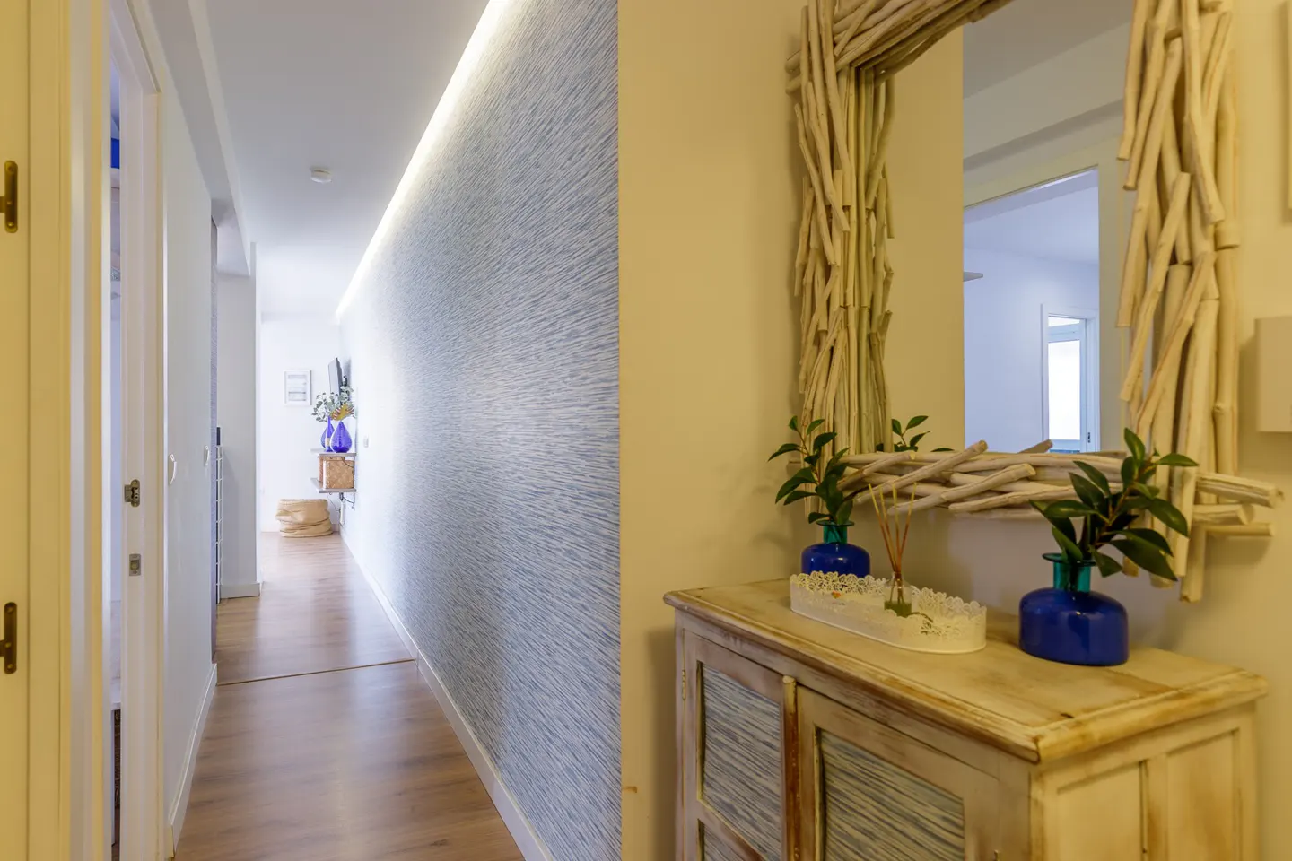 Hallway with wood floors, textured blue wallpaper, and a driftwood-framed mirror above a distressed cabinet with blue vases.