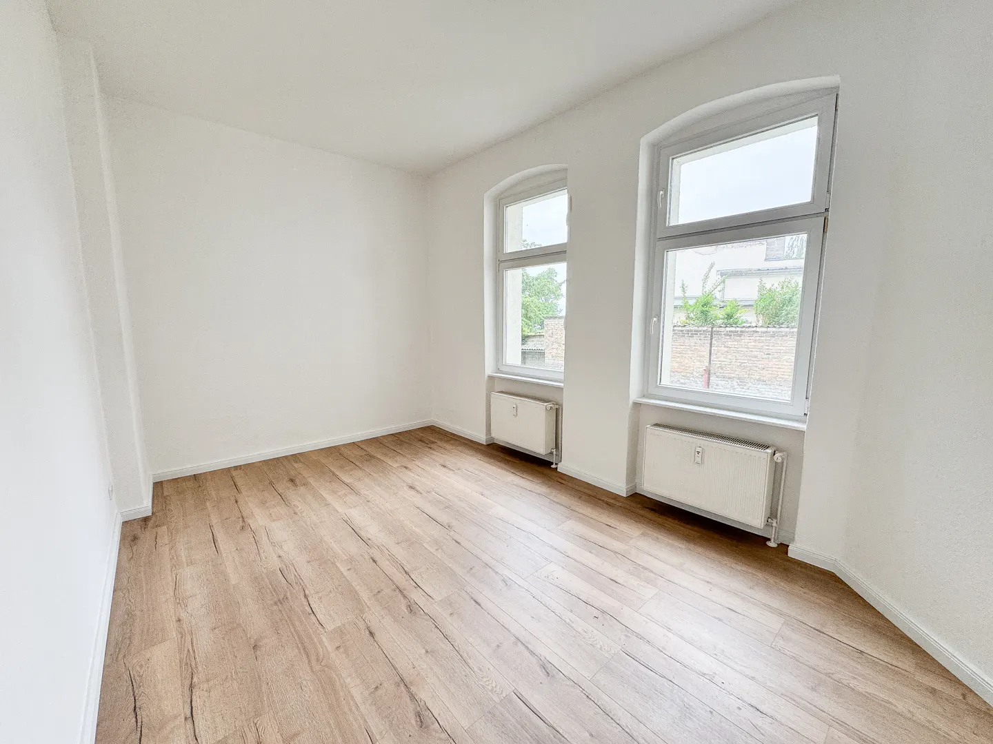 Bright, empty room with wood floors, white walls, two windows, and white radiators.