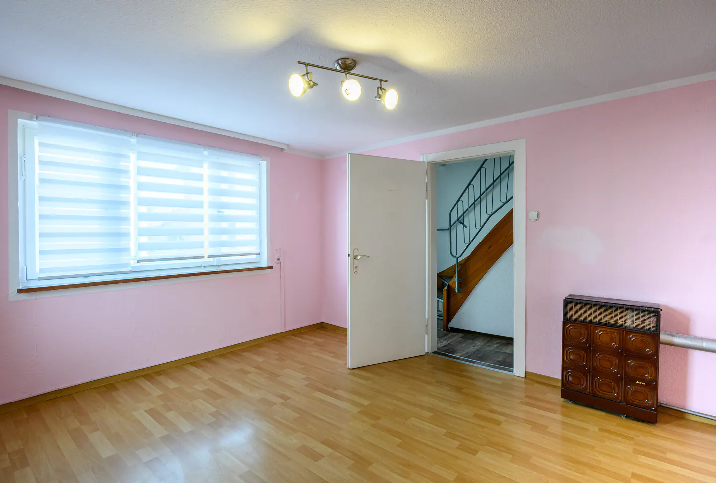 A room with pink walls, a wood floor, a window with blinds, and an open door to a staircase.