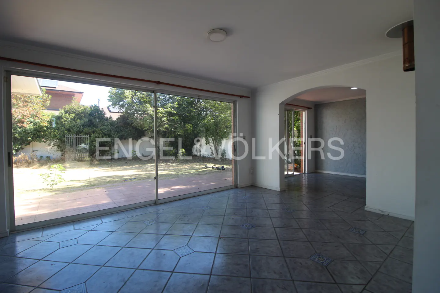 A bright, empty room with gray tile floors and a large sliding glass door to a green backyard. An arched doorway leads to another room.