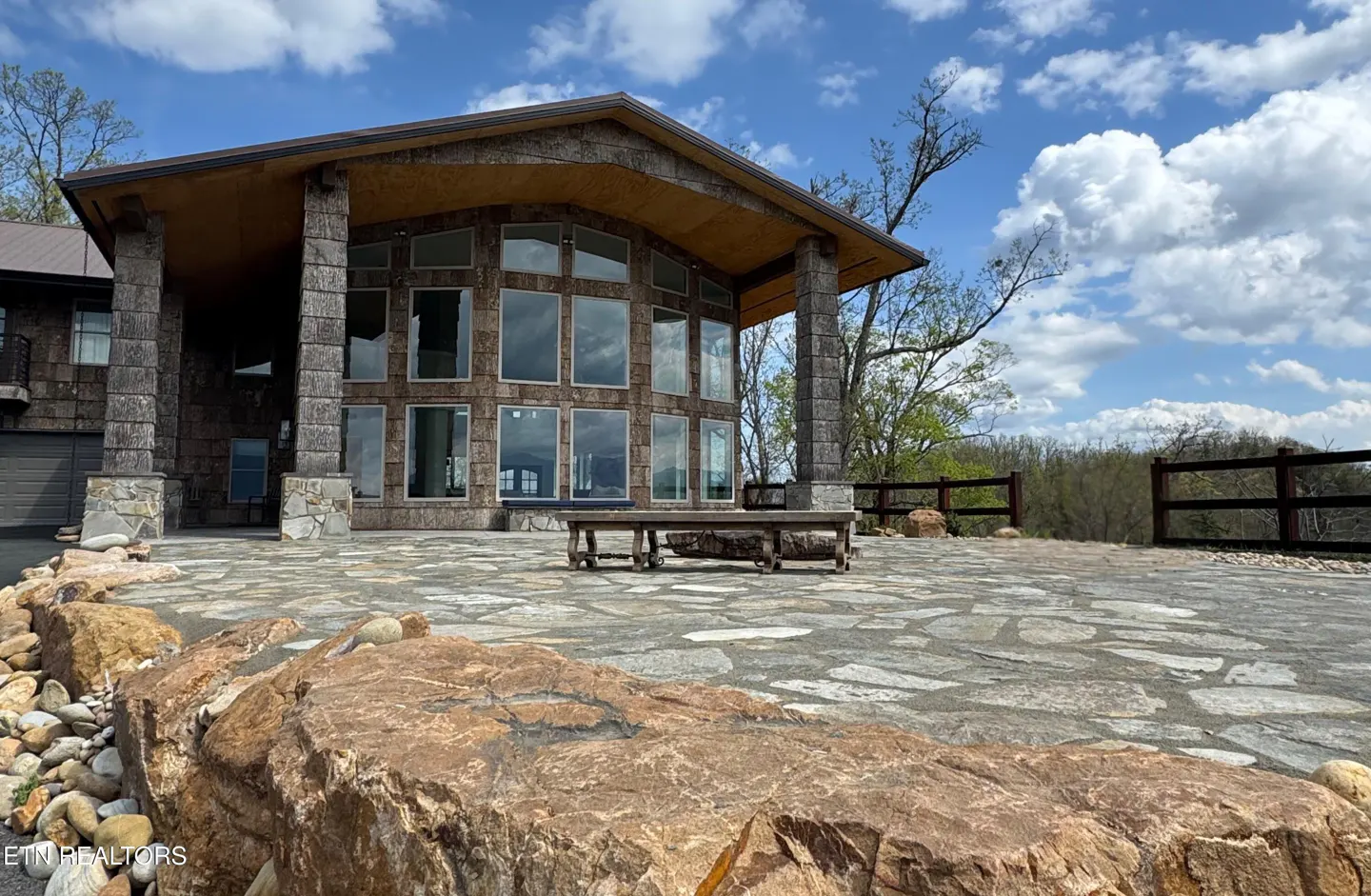 Exterior view of a stone house with a large patio, wooden bench, and a scenic view of trees and a blue sky.