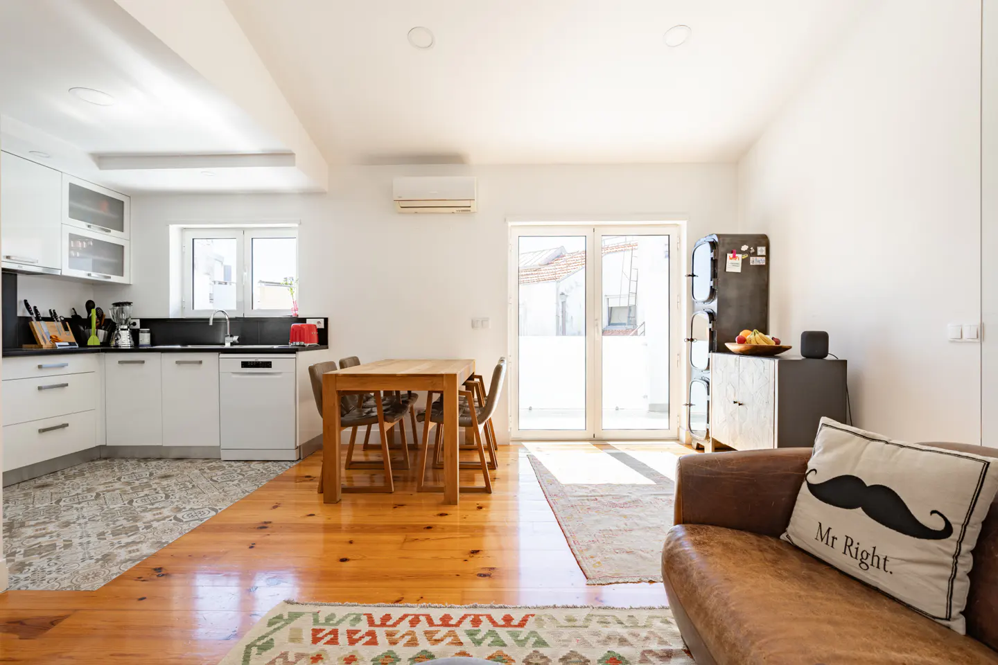 Bright open-plan kitchen and living area with wooden floors, a dining table, and a brown leather sofa with a mustache pillow.