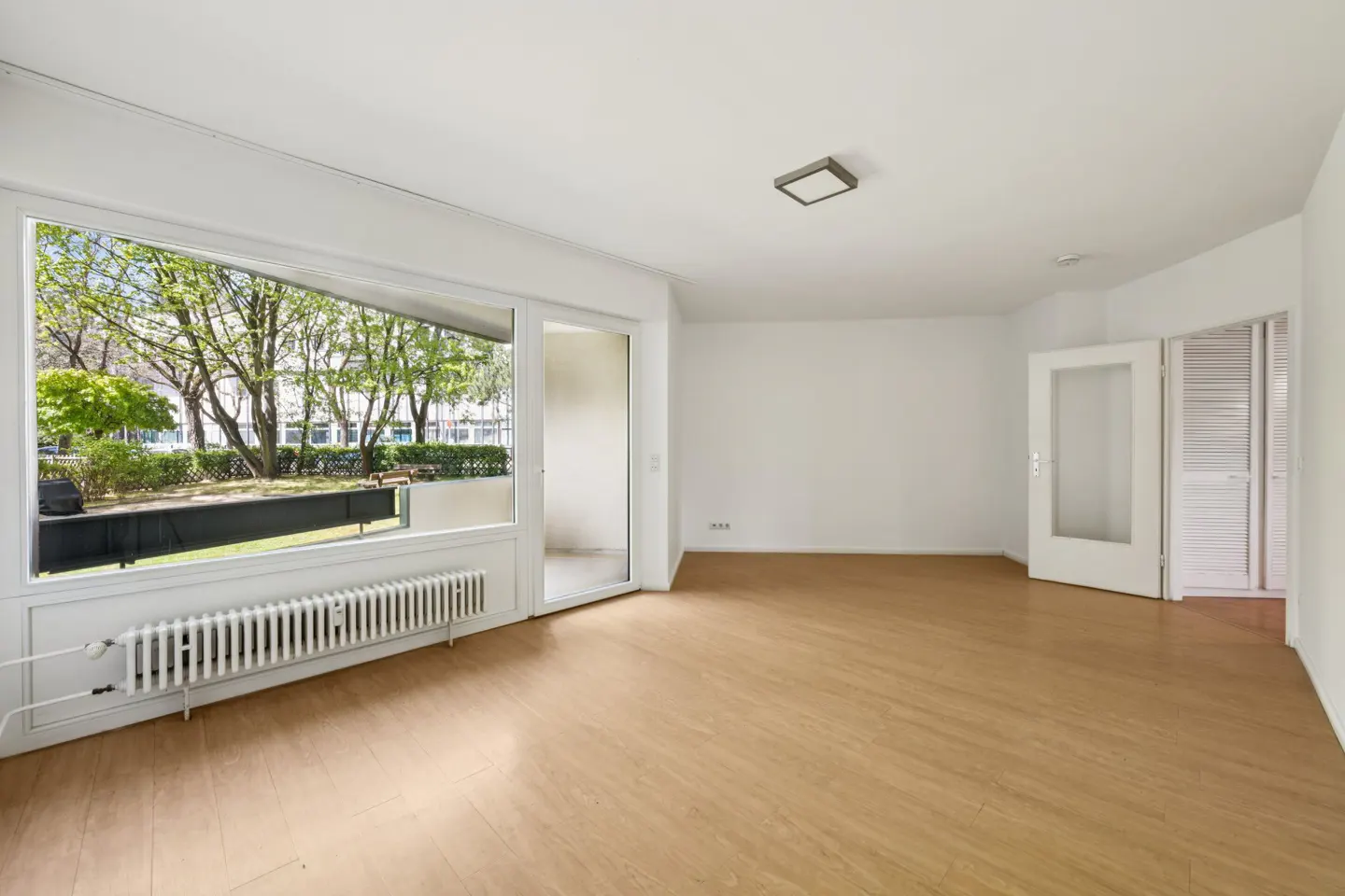 Bright, empty room with wood floors, white walls, and a large window overlooking a green outdoor space. A white radiator sits below the window.