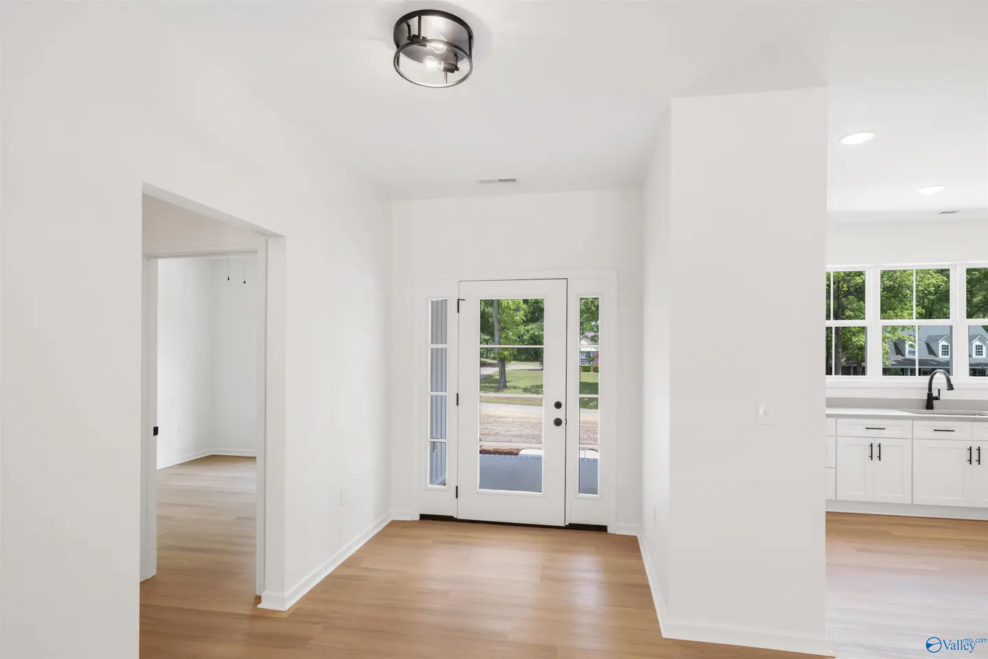 Bright foyer with white walls, wood floors, and a glass-paneled front door. A kitchen is visible to the right.