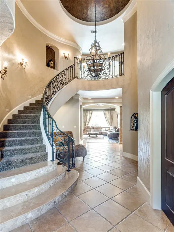Grand foyer with a curved staircase, wrought iron railing, and a large chandelier. Beige walls and tiled floors lead to a living room.