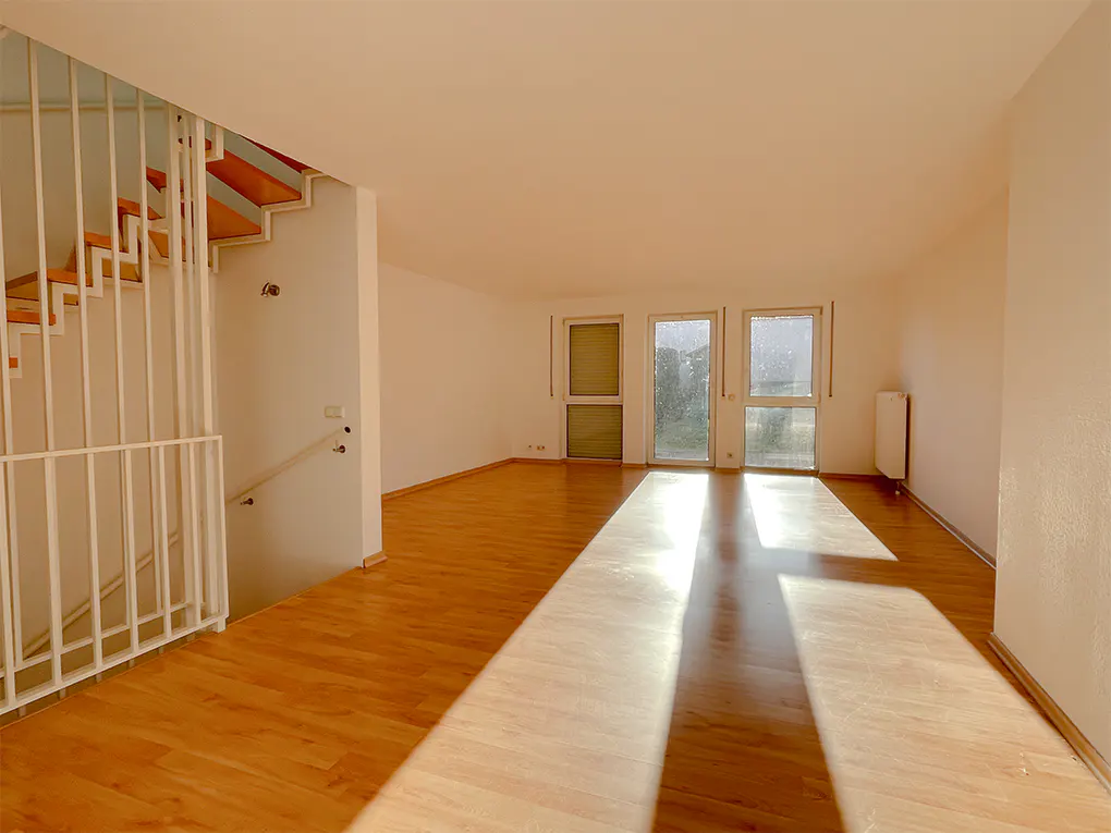 Bright, empty room with wood floors, white walls, and sunlight streaming through three windows. A white staircase is on the left.