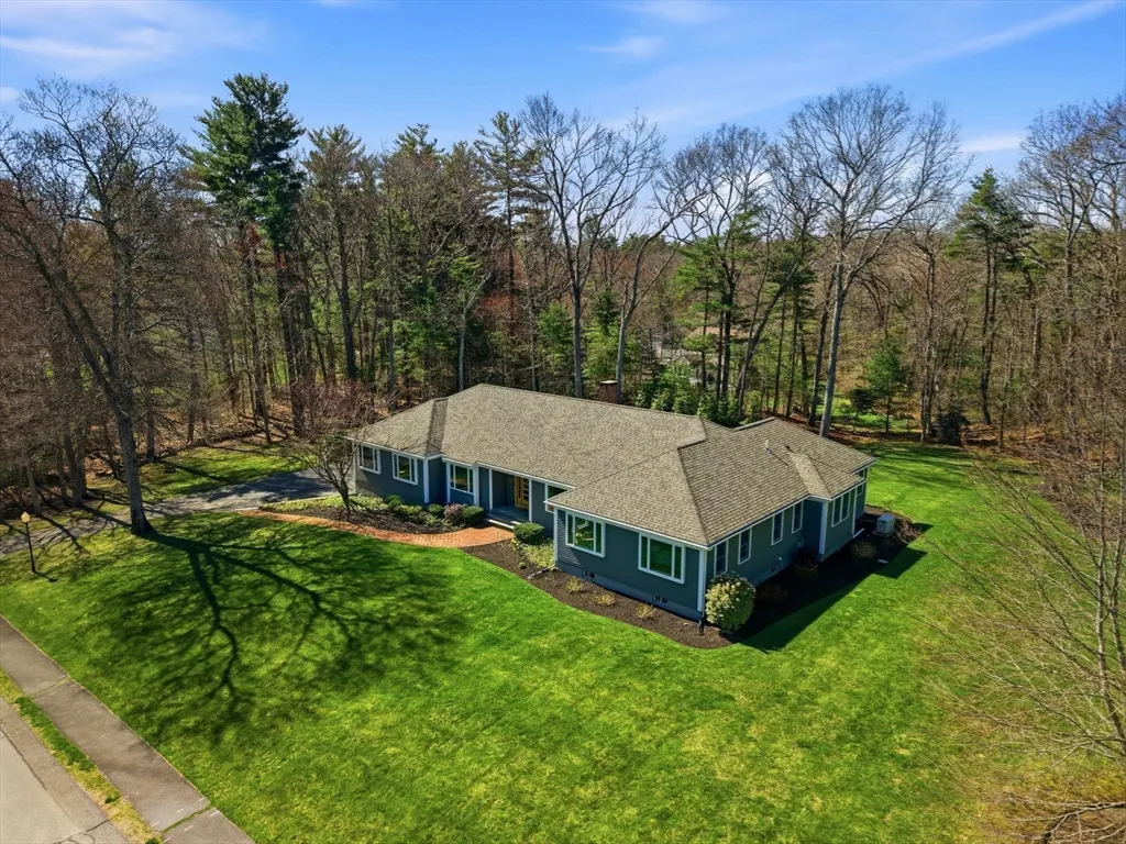 Aerial view of a grey ranch-style house with a shingled roof, surrounded by a large green lawn and tall, bare trees.